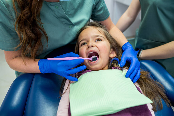malá holčička je ošetřena na malé zuby v zubním křesle kvalifikovaným pediatrem. Koncept stomatologické péče u batolat - Fotografie, Obrázek
