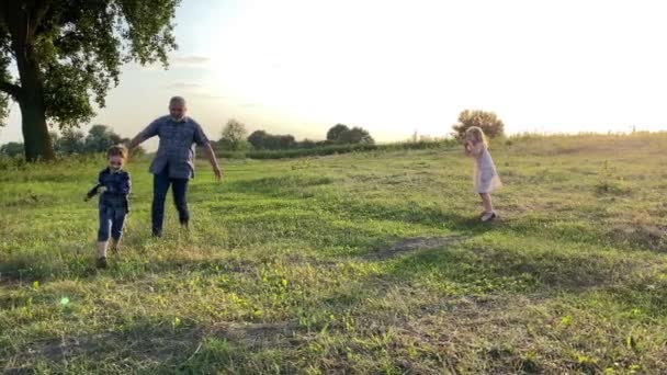 Cheerful father playing with kids at nature in fiery lights of sunset. Family with two children having leisure at meadow. Positive grandpa, little girl and boy enjoying spending time together outdoors - Séquence, vidéo