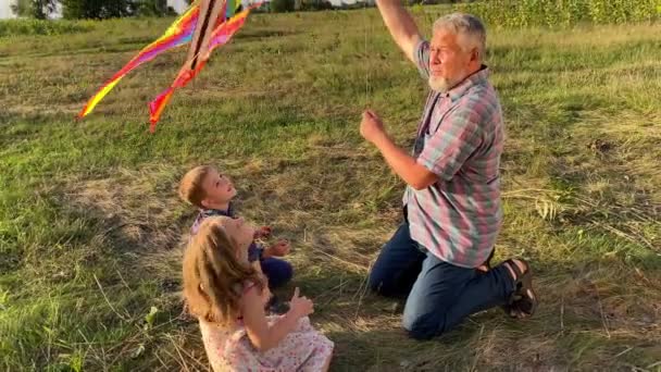 Charming kids with father preparing to fly kite at sunset outdoors. Family with two children spending time together at nature. Grandpa enjoying to have fun with kids at meadow. Happiness simple living - Séquence, vidéo