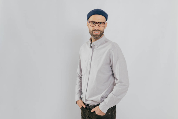Sideways shot of handsome pleased man wears stylish hat, keeps hands in pockets, stands against white background with copy space for your advertisement or promotion. Hipster guy models indoor - Photo, Image