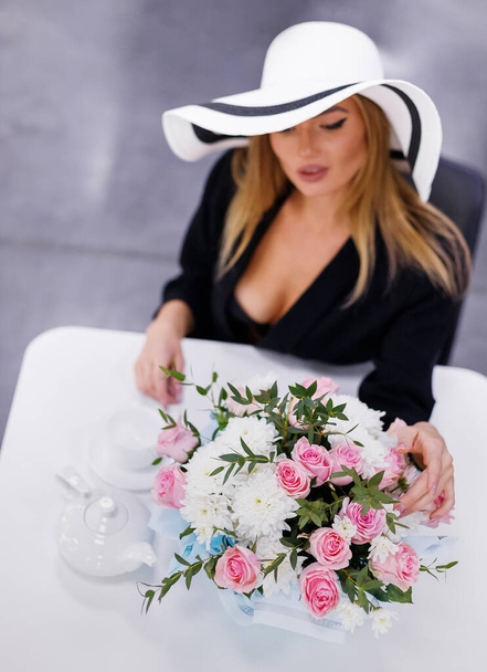 Bouquet of white chrysanthemums and pink roses on table in restaurant near glamorous woman. Top view of woman in white hat elegantly touches flowers sitting at table and drinking tea. Selective focus. - Foto, Imagem
