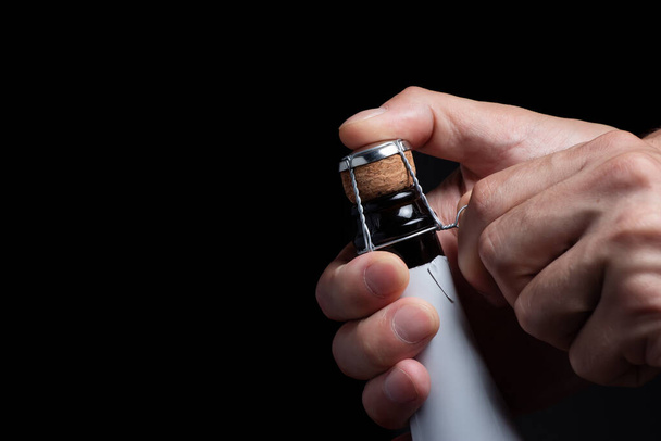Man's hand opens a bottle of champagne. The man unscrews the champagne muselet. Champagne bottle on a black background - Photo, Image