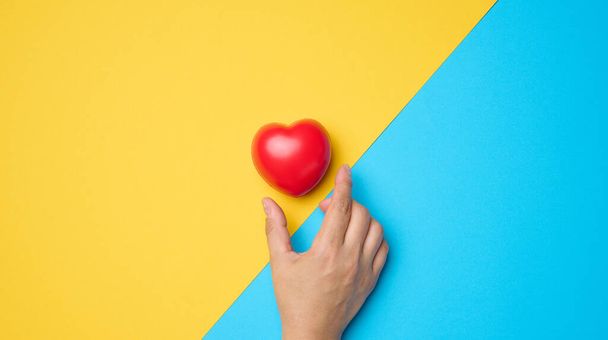 Red heart and a woman's hand reaches for it on a blue-yellow background - Foto, Bild