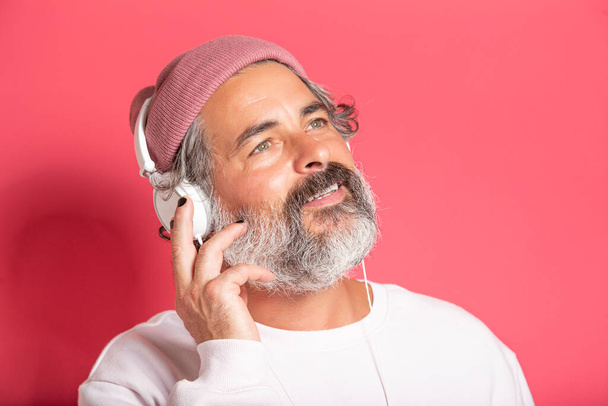Dark gray haired middle aged man 40s 50s in casual sweater listening to music with headphones isolated on pink background studio portrait - Valokuva, kuva