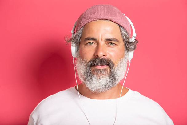 Close up of handsome grey-bearded man in earphones listening to music or talking on mobile phone in headphones, smiling happily at camera, standing over fuchsia background - Fotó, kép