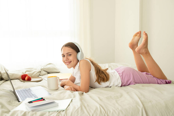 Iskoláslány készíti a házi feladatát, miközben otthon fekszik az ágyon, kezében egy csésze vízzel. Aranyos lány, 10 éves, fehér gyerek. Koncepciója virtuális iskola, tudás, távtanulás, online. - Fotó, kép
