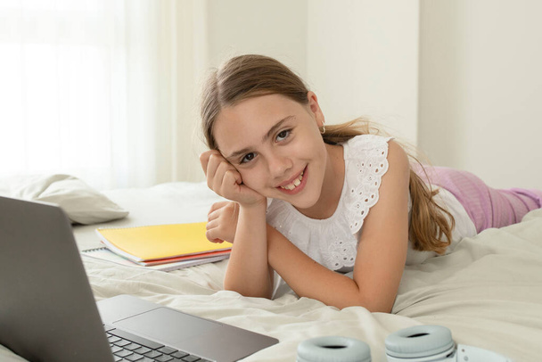10 yaşında beyaz bir kız, okul çocuğu evde yatakta ders çalışıyor. Mutlu çocuk dizüstü bilgisayarla ödev yapıyor. Evde eğitim kavramı, çocukluk, okula dönüş, web eğitimi, çevrimiçi sınıf, uzaklık öğrenimi - Fotoğraf, Görsel