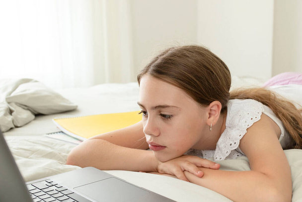 Upset schoolgirl in front of a laptop lying at home in the bedroom. Sad face. Girl is tired, bored listening to an online lesson. Concept of homeschooling, distance study, education, school kids. - 写真・画像