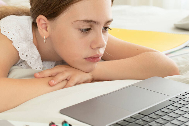 Upset schoolgirl in front of a laptop lying at home in the bedroom. Sad face. Girl is tired, bored listening to an online lesson. Concept of homeschooling, distance study, education, school kids. - Foto, imagen