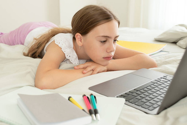 Feldúlt iskoláslány egy laptop előtt, aki otthon fekszik a hálószobában. Szomorú arc. A lány fáradt, unatkozik egy online leckét hallgatva. Fogalom az otthoni tanulás, távoktatás, oktatás, iskolás gyerekek. - Fotó, kép