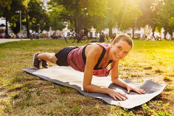 Happy girl of athletic build, with short hair, in sportswear for training, stands in plank on yoga mat in public park, in rays of sunlight. Outdoor workout concept, active lifestyle - Photo, Image