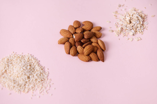 Selective focus on a handful of raw organic almond nuts, near scattered sesame seeds and oat-flakes on a pink background, copy advertising space. View from above. Flat lay. Still life. Superfood. - Foto, afbeelding