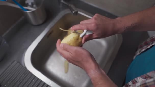 Bachelor is cooking dinner at home in kitchen. Healthy food theme, man peels the skin on raw potato in close-up over kitchen sink. Non GMO vegetables from the farm. Mans hands peeling potato peels.  - Materiaali, video