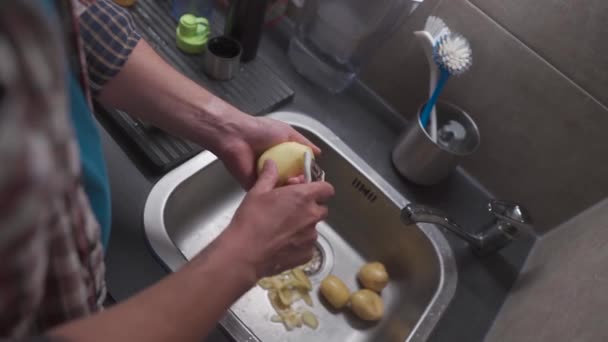 Bachelor is cooking dinner at home in kitchen. Healthy food theme, man peels the skin on raw potato in close-up over kitchen sink. Non GMO vegetables from the farm. Mans hands peeling potato peels.  - Séquence, vidéo