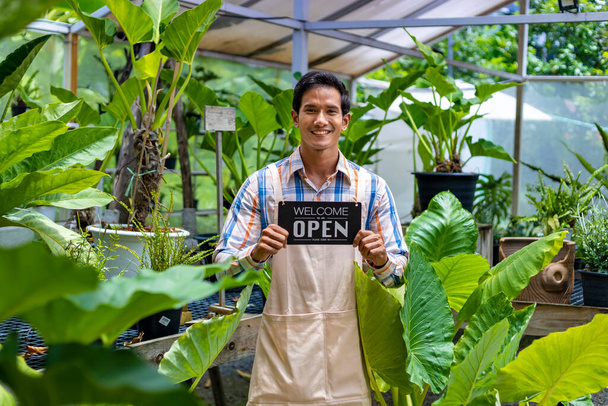 Asian gardener is holding opening sign to welcome customer to their tropical nursery plant center full of exotic fern species - Photo, image