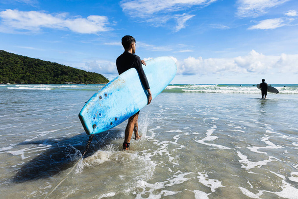 Young fit and firm body teenage guy walking into sea holding soft board ready for extreme sports surfing on beach holidays on clear day, blue skies. Concept for teen hobby and adventure in vacation. - Fotó, kép