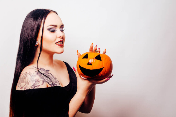 Piękna brunetka trzyma dynię w rękach i patrzy na nią. Kobieta z makijażem na Halloween, ciemna szminka. Model w kolorze białym - Zdjęcie, obraz