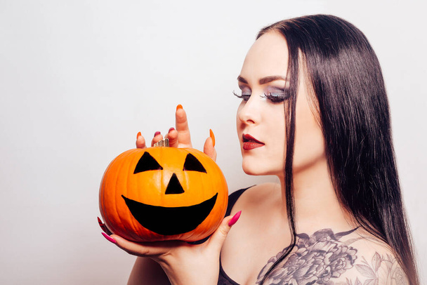 Una hermosa morena sostiene una calabaza en sus manos y la mira. Mujer con maquillaje de Halloween, lápiz labial oscuro. Modelo en blanco - Foto, imagen