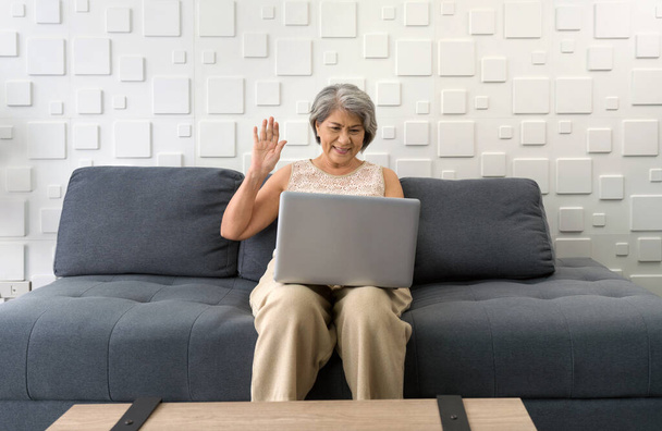 Asian gray hair old woman waving hand via remote communication through laptop computer in the living room.  - Photo, Image