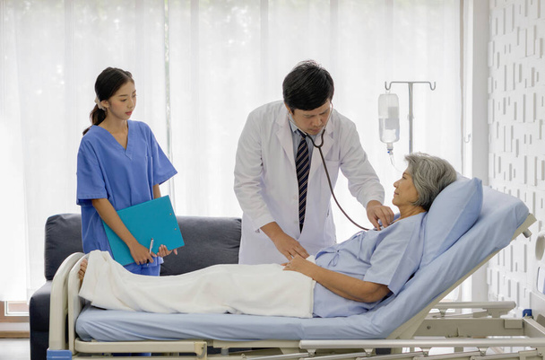 Senior female patient and doctor. Asian doctor use a stethoscope to check the heartbeat of the elderly patient. Young nurse stand next to the bed, holding document folder. - Foto, imagen