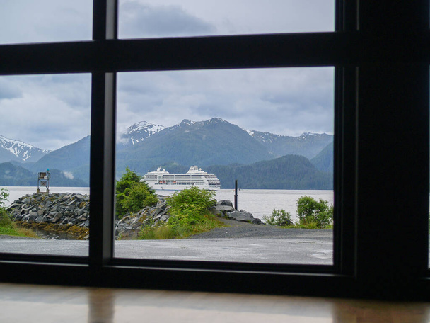 View through window of dramatic Alaskan scenery and passing cruise-ship under cloudy sky. - Zdjęcie, obraz