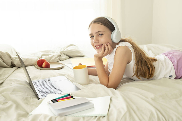 10-jarig meisje lachend met laptop in bed, theekopje vasthoudend, naast bord fruit, notitieboekjes. Koptelefoon op het hoofd van het kind. Concept van comfortabele studie, thuisschool, online onderwijs - Foto, afbeelding