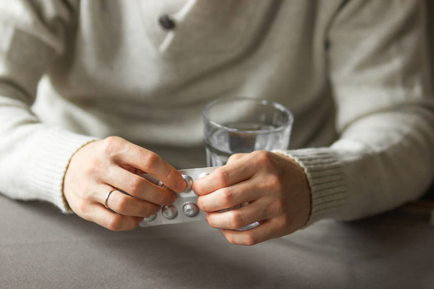 Mans hands holds and open silver blister with pills, and ready to take medicines with glass of water. Sick man need medicine from headache and cold, painkiller at home. Health care concept. - 写真・画像