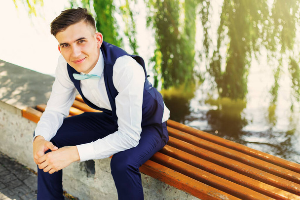 A stylish groom sits on a bench near the lake and looks at the camera - 写真・画像