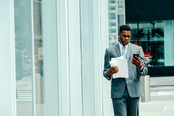 Young adult businessman using smartphone outside wearing suit, tie and headphones holding documents - 写真・画像