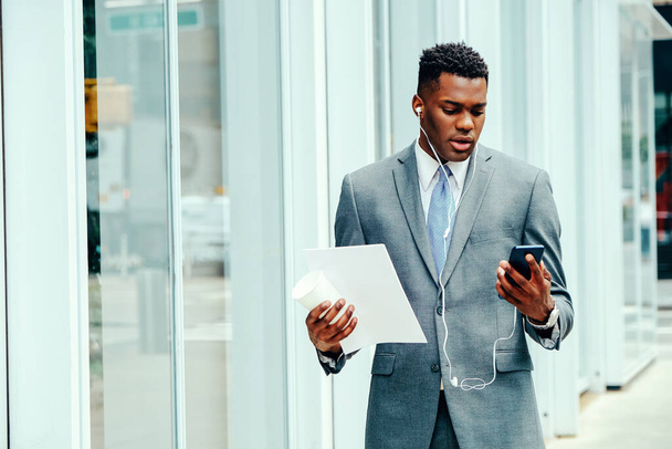 Young adult entrepreneur using smartphone outside wearing suit, tie and headphones holding documents - Фото, изображение