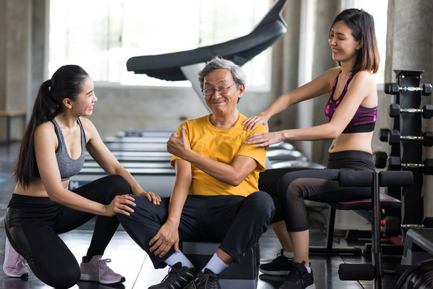 Smiling old man thumb up after getting massage by beautiful Asian women. Relief after treadmill cardio exercise at fitness gym. Happy healthy lifestyle for elderly senior guy or pensioner. - Photo, Image