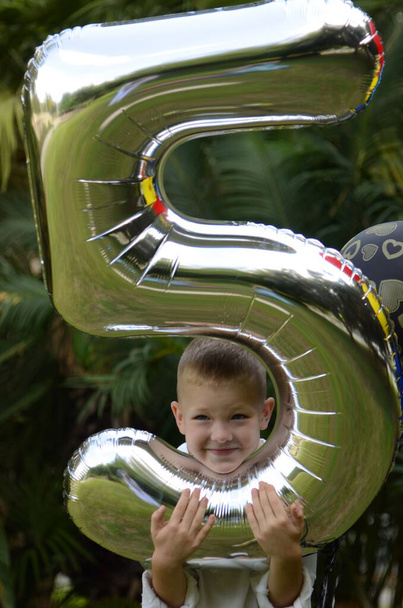 O menino comemora seu aniversário. Idade cinco anos. Criança com balões no parque. Férias infantis, presentes, bom humor.  - Foto, Imagem