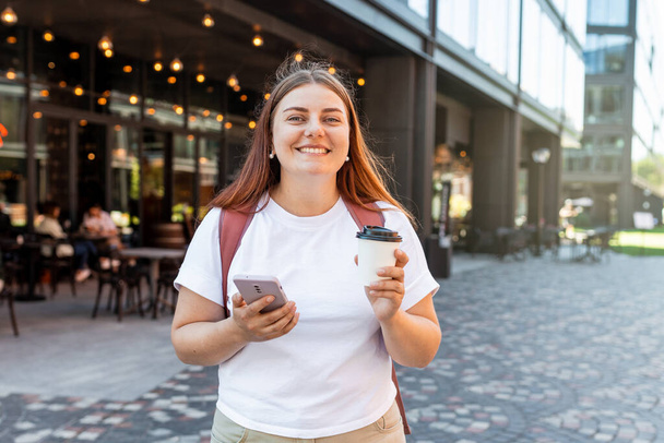 Kuva nuori nainen ulkona kadulla etsivät syrjään chattailuun matkapuhelimella kahvin juominen. Nuoret naiset käyttävät älypuhelinta ja juovat teetä kaupungissa. Kaupunkien elämäntavan käsite. - Valokuva, kuva