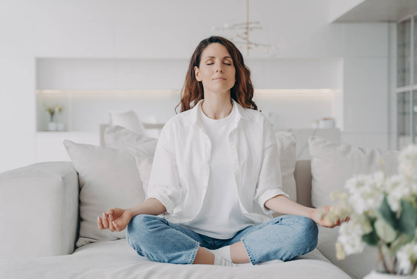 Serene hispanic woman practicing meditation on comfortable sofa at home. Calm female sitting in lotus position on couch, doing yoga, breathing fresh air, relaxing for stress relief. - Photo, image