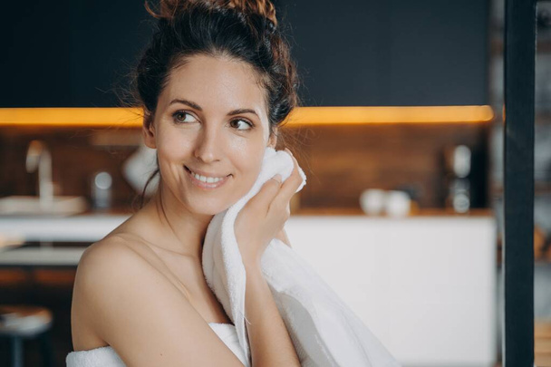 Smiling hispanic woman wiping face with white cotton towel after shower, happy pretty female with naked shoulder enjoying her fresh glowing skin after skincare beauty morning routine at home. - Foto, afbeelding