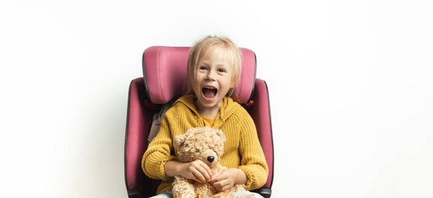 Cute little caucasian girl fastened with a belt in a safety car seat isolated on white background. Kid safety while traveling by car. Happy toddler girl holding teddy bear. Adventure concept - Foto, Imagen