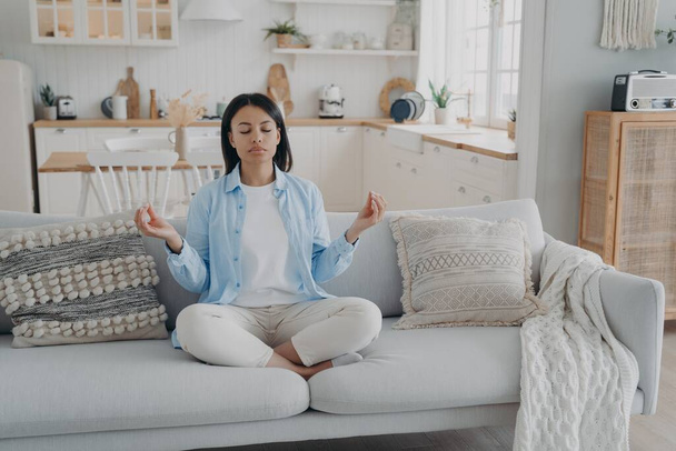 Zen, tranquility and breath control. Spanish woman is practicing yoga gymnastics in living room. Girl sitting on couch in lotus pose and meditating with her eyes closed. Relaxation and stress relief. - Photo, image