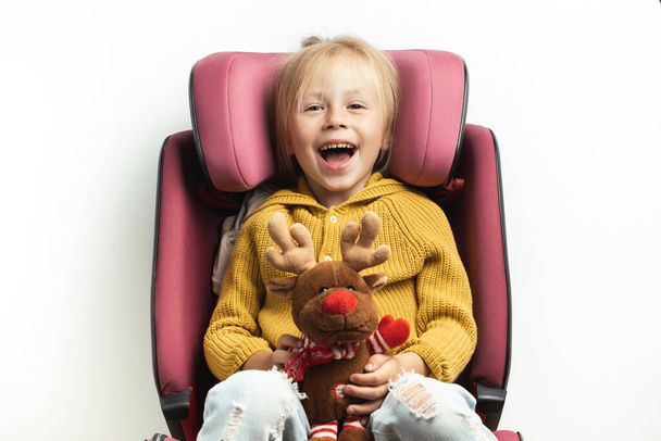 Cute little girl holding stuffed Rudolph Red-Nosed Reindeer toy while sitting in a safety car seat. Kid safety while traveling by car. Isolated on white. Merry Christmas. Holiday adventure concept - Foto, Imagen