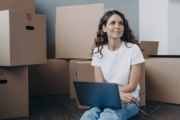 Pensive spanish woman with laptop sit on floor near heap of cardboard boxes, search moving company on internet, buy furniture for home online, dreaming about new home on relocation day. - Foto, Imagem