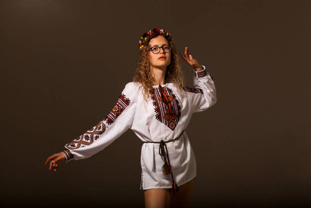A model in national dress poses on a dark background. A young woman with curly hair and a wreath of flowers on her head. The model's legs are bare and the shirt is tied with a rope. Eco style of clothing - Zdjęcie, obraz