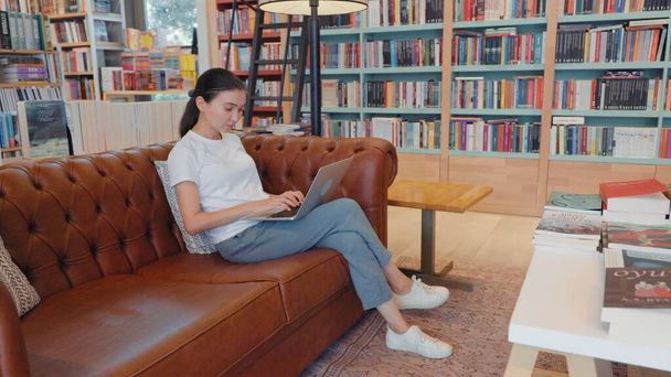 Young woman texting on laptop sitting on sofa in library, office, home. High quality 4k footage - Foto, imagen