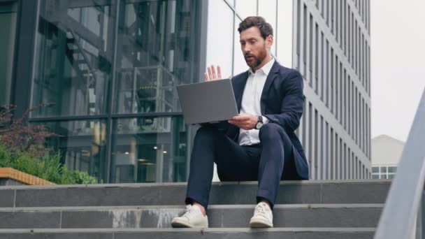 Successful businessman talking on video call on laptop negotiating conducting interview outdoors via online conference young male professional worker sitting on street chatting on webcam waving hello - Video, Çekim