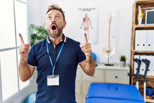 Hombre fisioterapeuta de mediana edad que trabaja en la clínica de recuperación del dolor asombrado y sorprendido mirando hacia arriba y señalando con los dedos y los brazos levantados.  - Foto, imagen