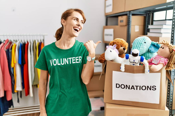 Bella donna caucasica che indossa la t-shirt volontaria alle donazioni stand sorridente con volto felice guardando e indicando il lato con pollice in su.  - Foto, immagini
