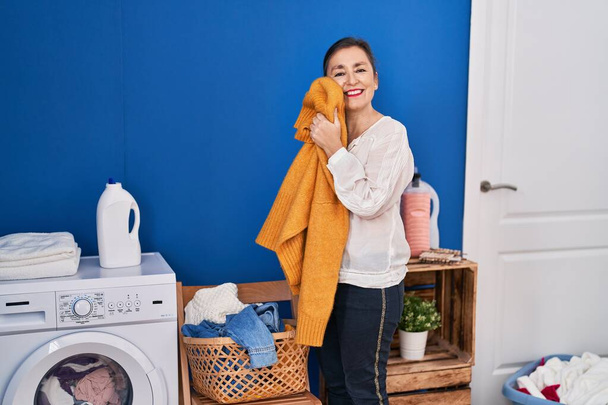 Middle age woman washing clothes touching softener sweater with face at laundry room - Φωτογραφία, εικόνα