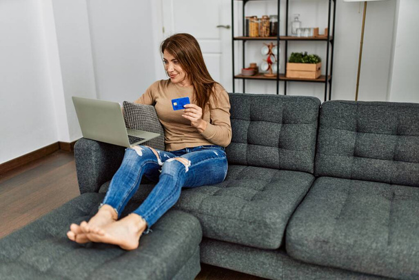 Mulher caucasiana de meia idade usando smartphone e cartão de crédito sentado no sofá em casa. - Foto, Imagem