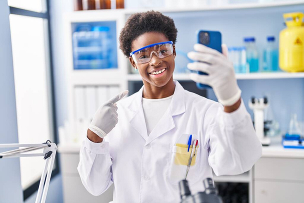 Mujer afroamericana trabajando en laboratorio científico haciendo videollamada con smartphone sonriendo feliz señalando con la mano y el dedo  - Foto, Imagen