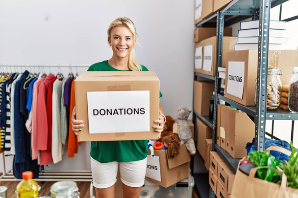 Молода блондинка-волонтерка посміхається щасливо тримає коробку пожертвувань у благодійному центрі
. - Фото, зображення