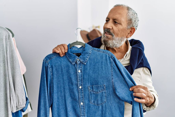 Hombre mayor de pelo gris sonriendo confiado sosteniendo la camisa en la tienda de ropa - Foto, imagen