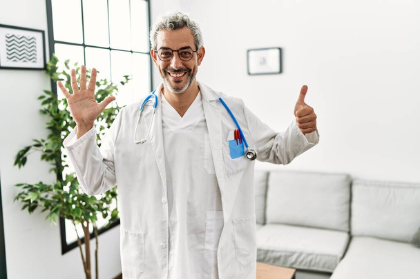 Latynos w średnim wieku, noszący mundur lekarza i stetoskop w poczekalni, pokazujący i wskazujący palcami numer sześć, uśmiechający się z pewnością siebie i szczęściem..  - Zdjęcie, obraz